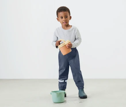 Un jeune garçon tient un seau orange dans ses mains, portant un pantalon de pluie bleu et des bottes en caoutchouc.
