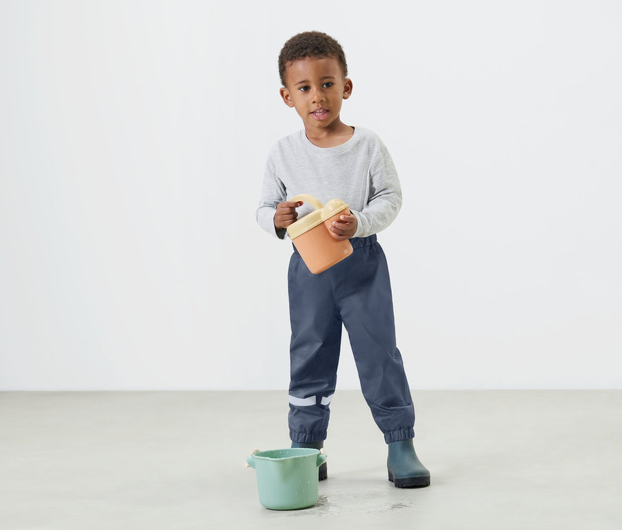 Un jeune garçon tient un seau orange dans ses mains, portant un pantalon de pluie bleu et des bottes en caoutchouc.