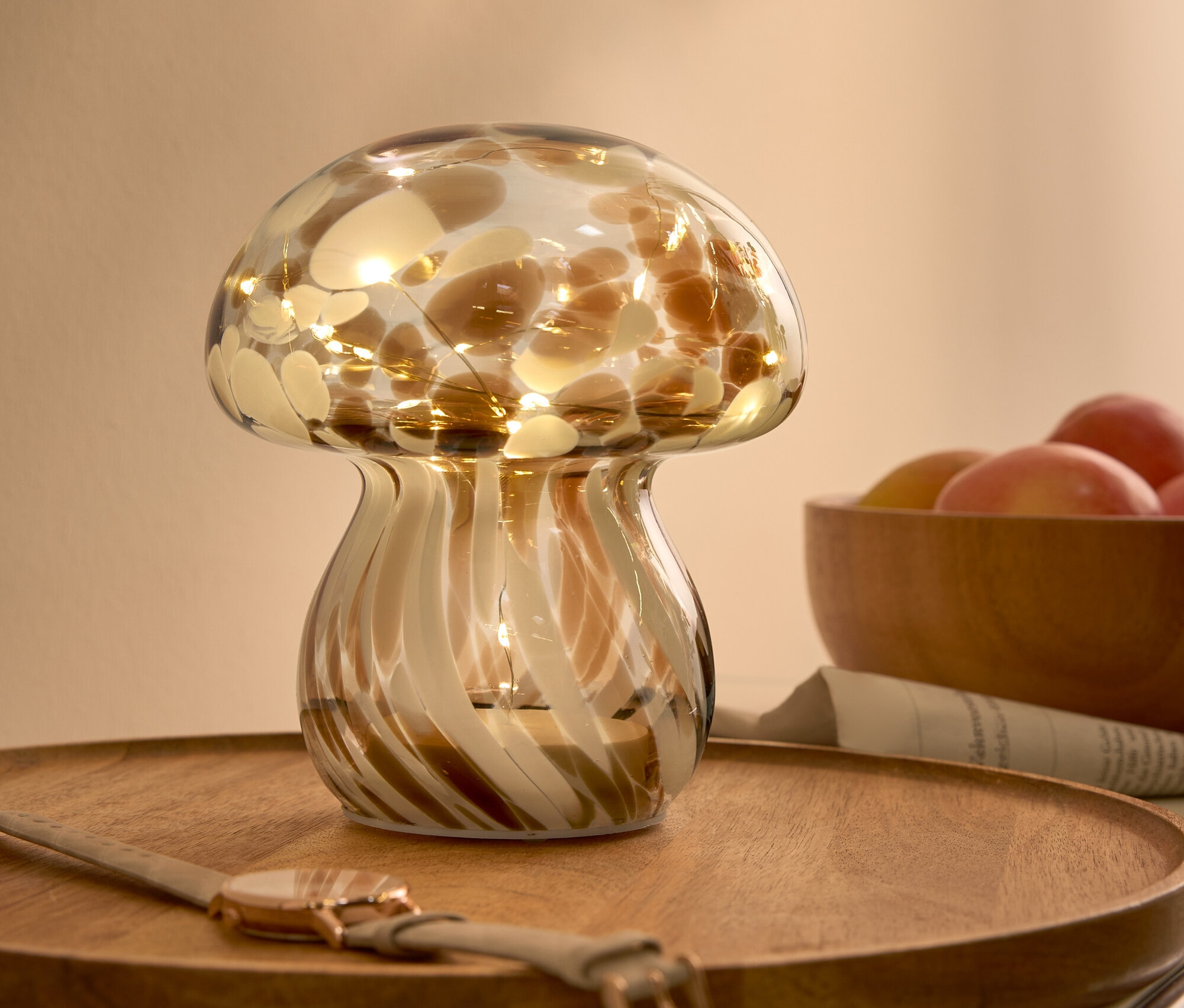 Lampe décorative en verre en forme de champignon avec des taches brunes et des lumières, sur un plateau en bois à côté d'une montre et d'un bol de fruits.