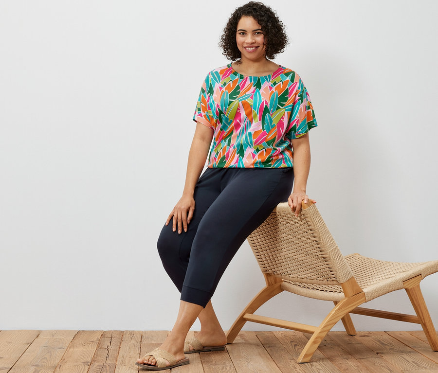 Une femme est assise sur une chaise en osier, portant un pantalon 3/4 en jersey bleu foncé et un chemisier à encolure dégagée à imprimé intégral.