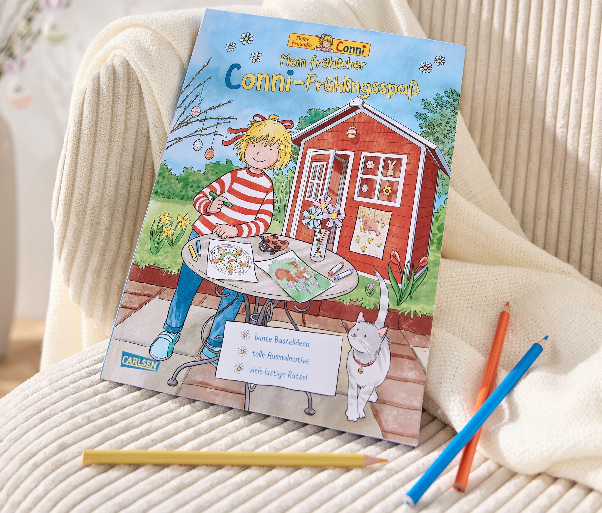 Le livre « Mon joyeux printemps avec Conni » est posé sur un fauteuil couleur crème à côté de crayons de couleur.