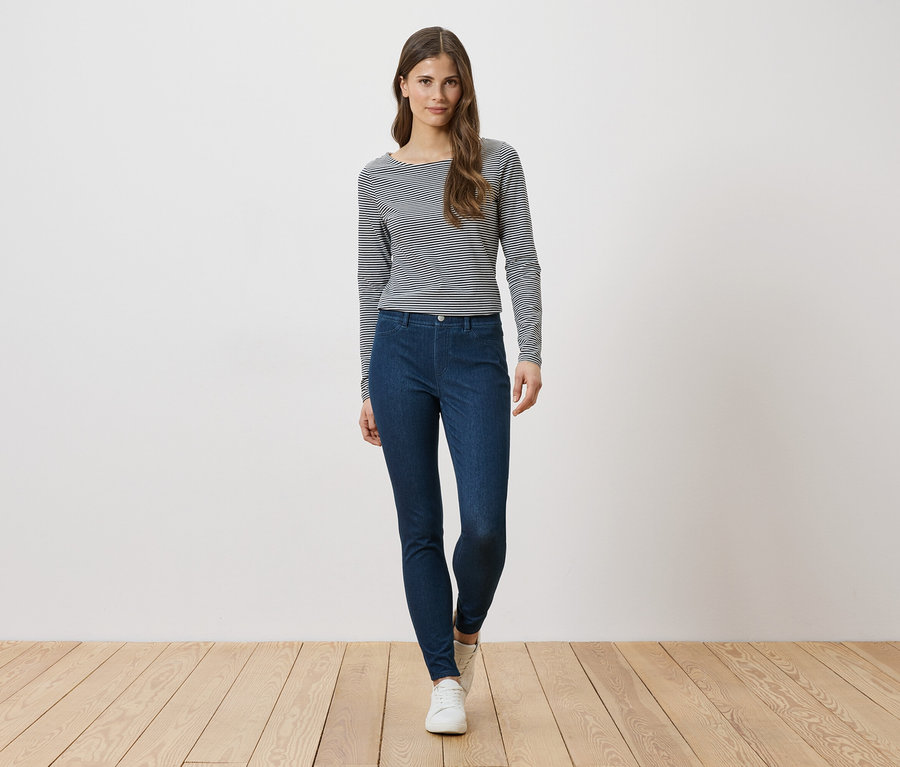 Femme portant un haut rayé à manches longues et un jean bleu, debout devant un mur blanc.