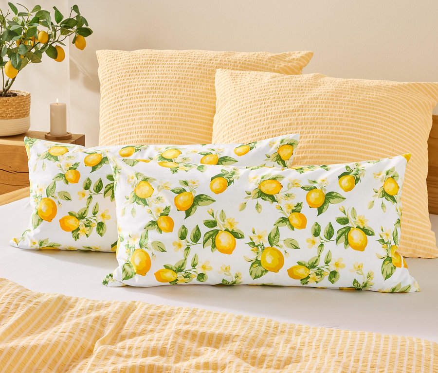 Sur un lit, deux coussins à motifs de citrons sont placés devant deux coussins jaunes. À gauche, un citronnier est posé à côté d'une bougie.
