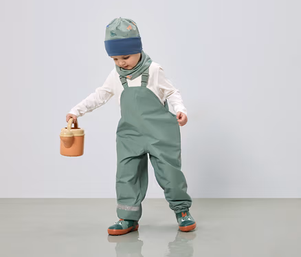 Un enfant portant une salopette verte, un bonnet et un foulard à motif de dinosaures tient un arrosoir.