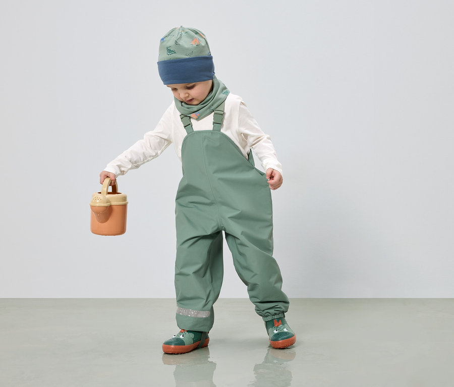 Un enfant portant une salopette verte, un bonnet et un foulard à motif de dinosaures tient un arrosoir.
