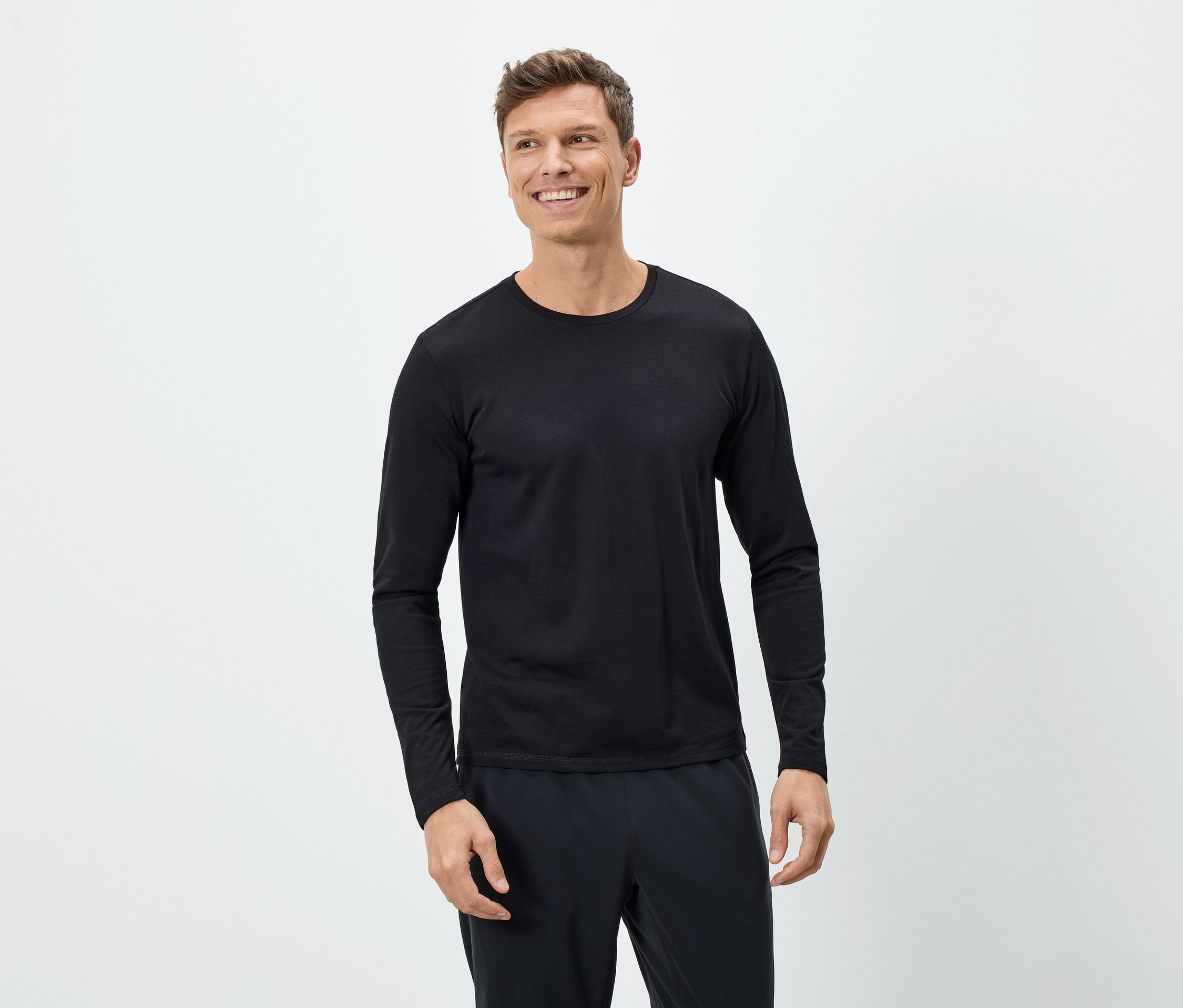 Un homme souriant porte un T-shirt noir à manches longues et un pantalon noir sur un fond blanc.