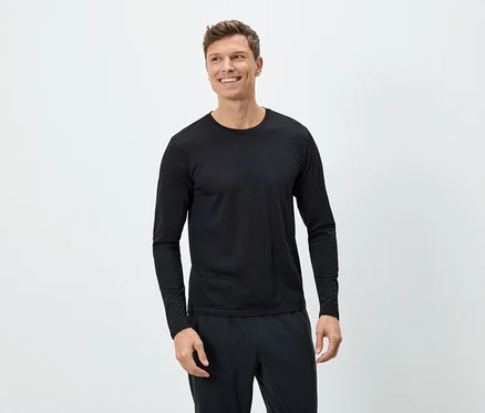 Un homme souriant porte un T-shirt noir à manches longues et un pantalon noir sur un fond blanc.
