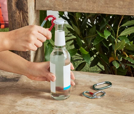 Des mains ouvrent une bouteille en verre transparent avec un mousqueton rouge sur un banc en bois, avec deux autres mousquetons à côté.