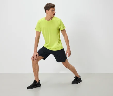 Un homme en tenue de sport s'étire. Il porte un t-shirt vert néon, un short noir et des baskets noires.