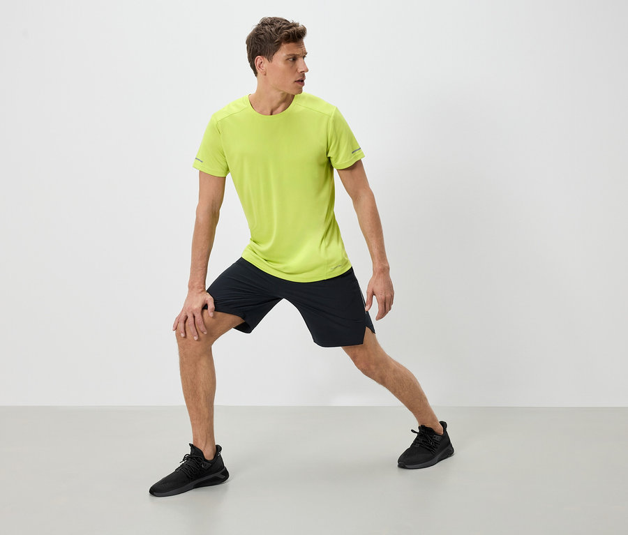 Un homme en tenue de sport s'étire. Il porte un t-shirt vert néon, un short noir et des baskets noires.