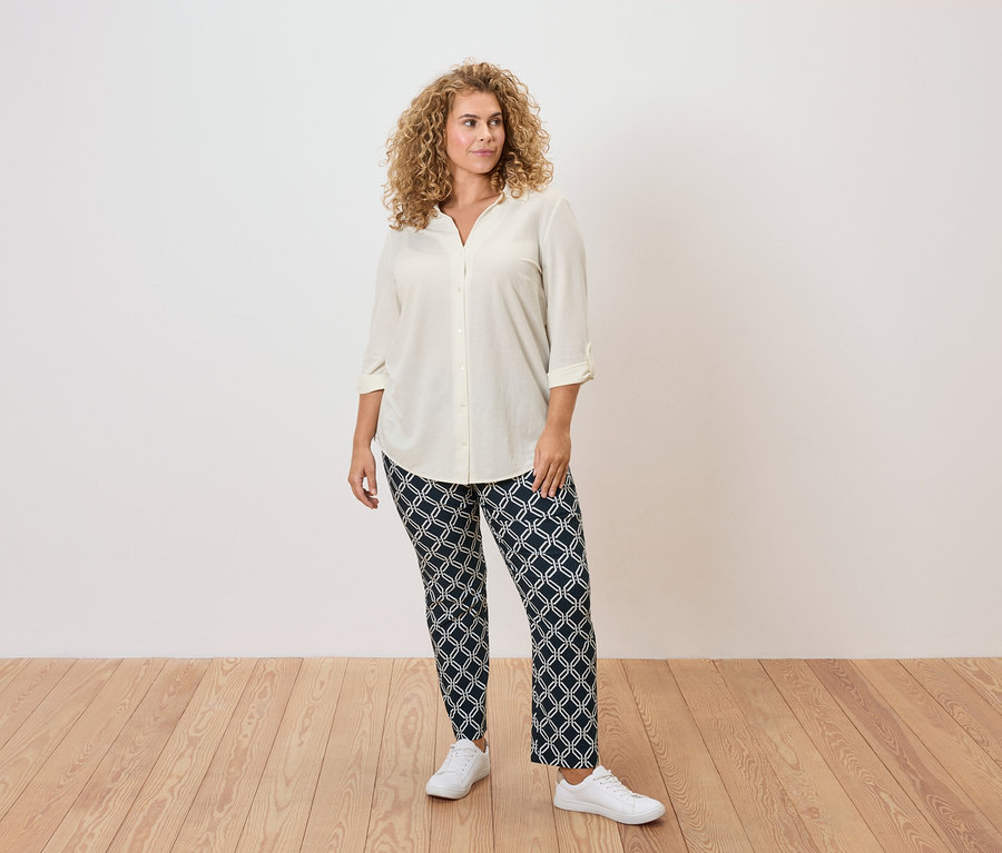 Une femme aux cheveux bouclés porte un chemisier crème, un pantalon à motifs et des baskets blanches.