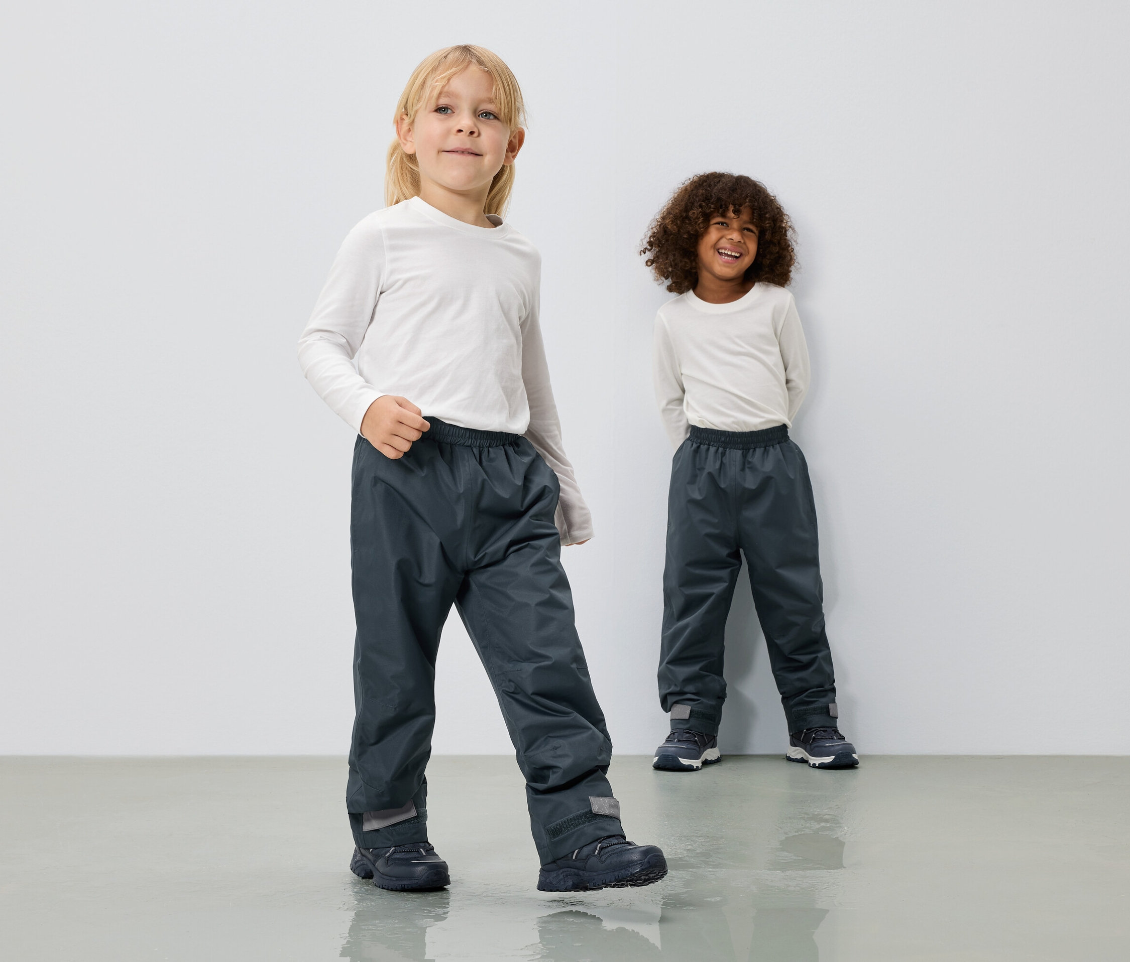 Deux enfants portent de longs pantalons de pluie gris et des hauts blancs à manches longues devant un mur blanc.