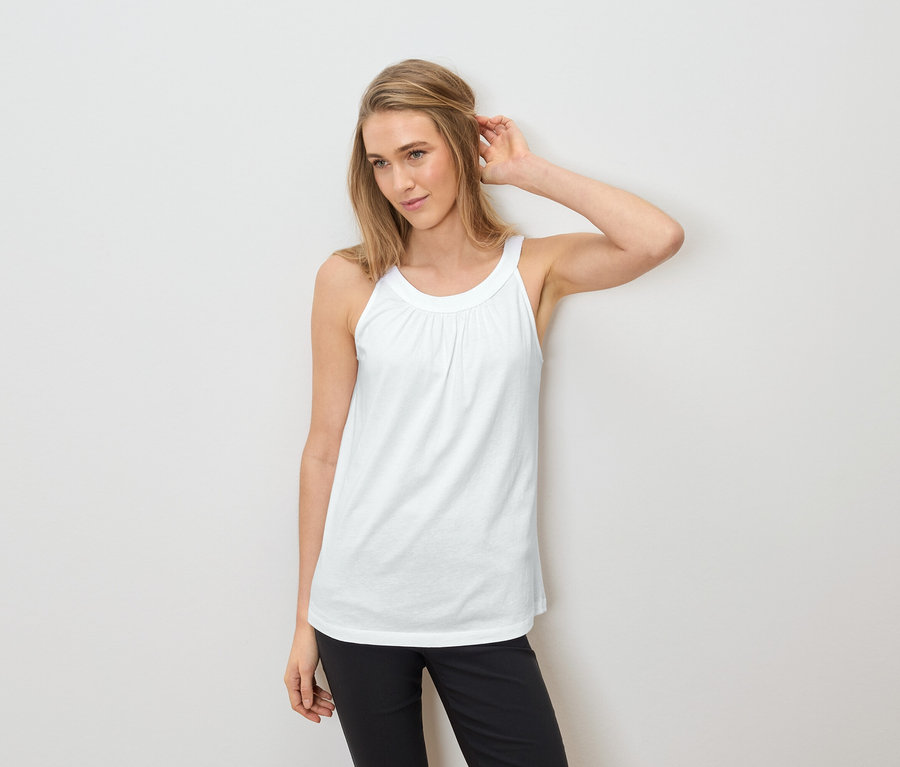 Femme portant un débardeur blanc et un pantalon noir, une main dans les cheveux.