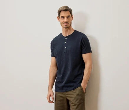 Homme portant un t-shirt Henley bleu et un pantalon kaki posant contre un mur blanc.