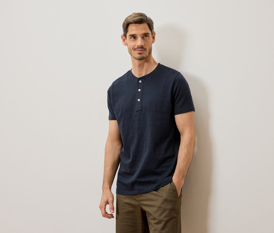 Homme portant un t-shirt Henley bleu et un pantalon kaki posant contre un mur blanc.