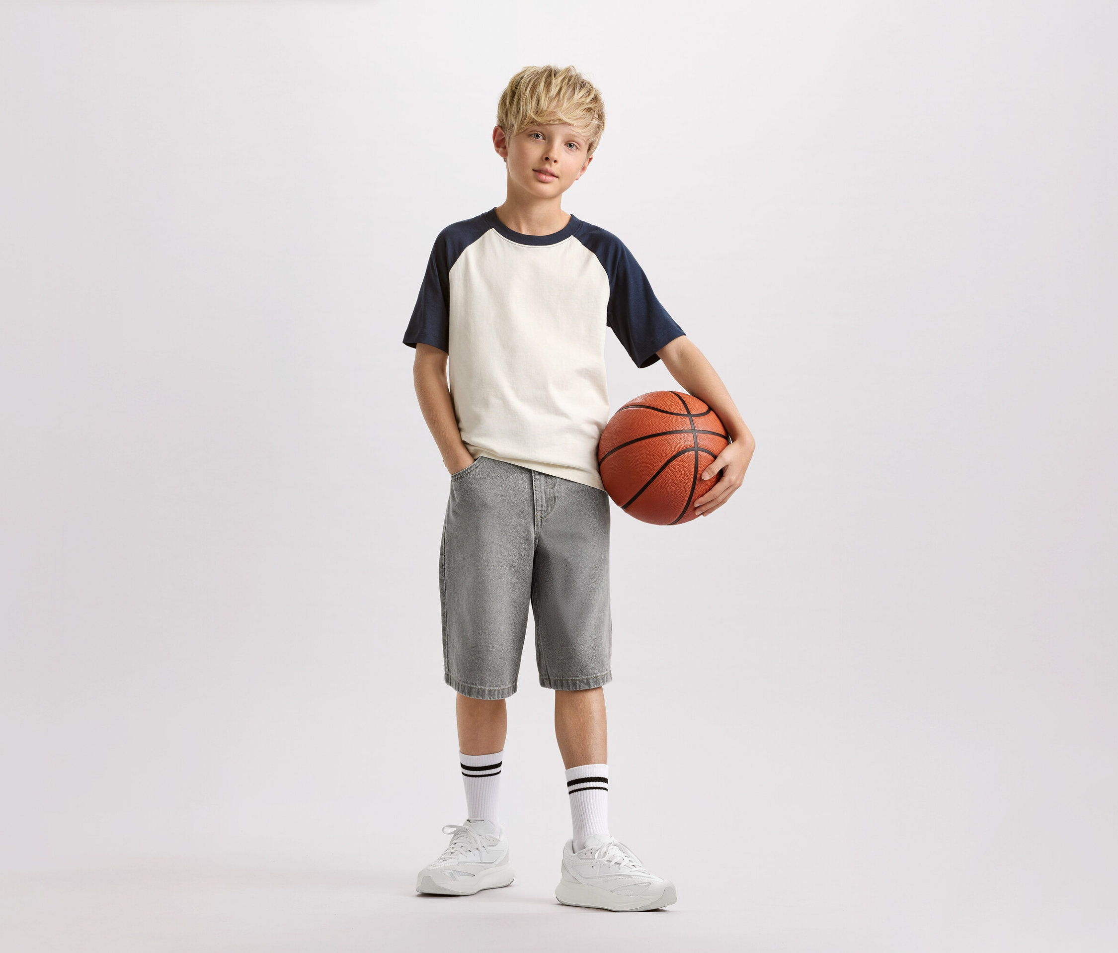 Un garçon aux cheveux blonds tient un ballon de basket, porte un t-shirt clair à manches bleu marine, un short gris et des chaussettes blanches à rayures noires.