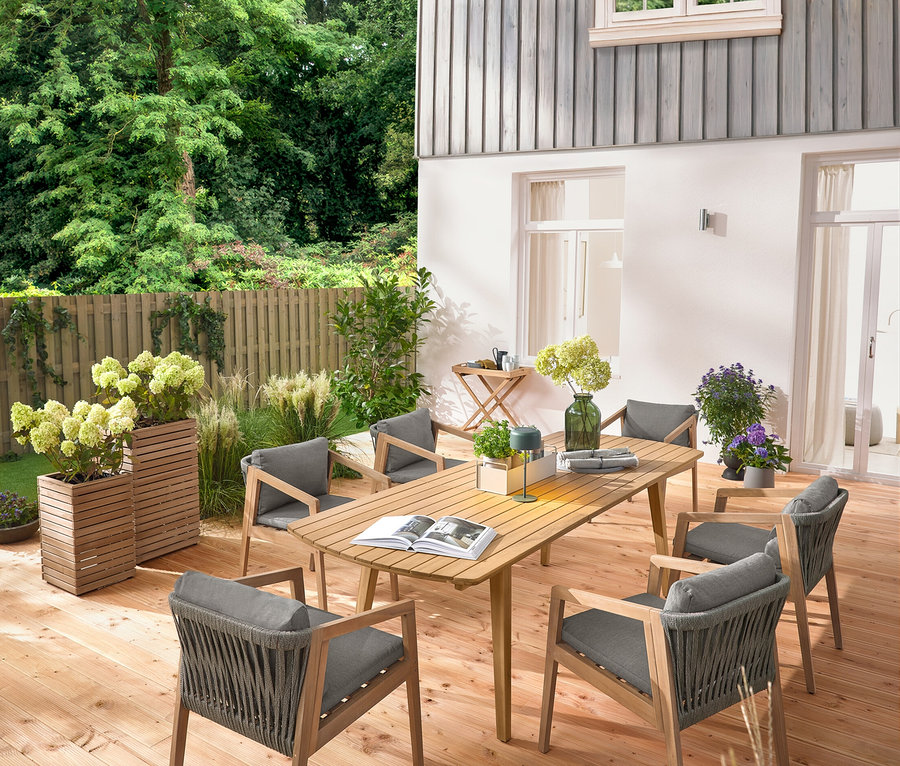 Sur une terrasse en bois se trouvent une table de jardin à rallonges avec façades arrondies, env. 180-240 cm, six chaises de jardin classiques, une tablette de jardin, une lampe de table solaire à batterie, vert jungle et 2 colonnes à plantes « Tinus ».