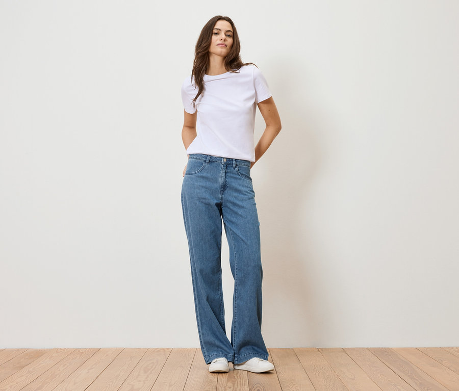 Une femme se tient devant un mur blanc, portant un t-shirt blanc et un jean bleu.