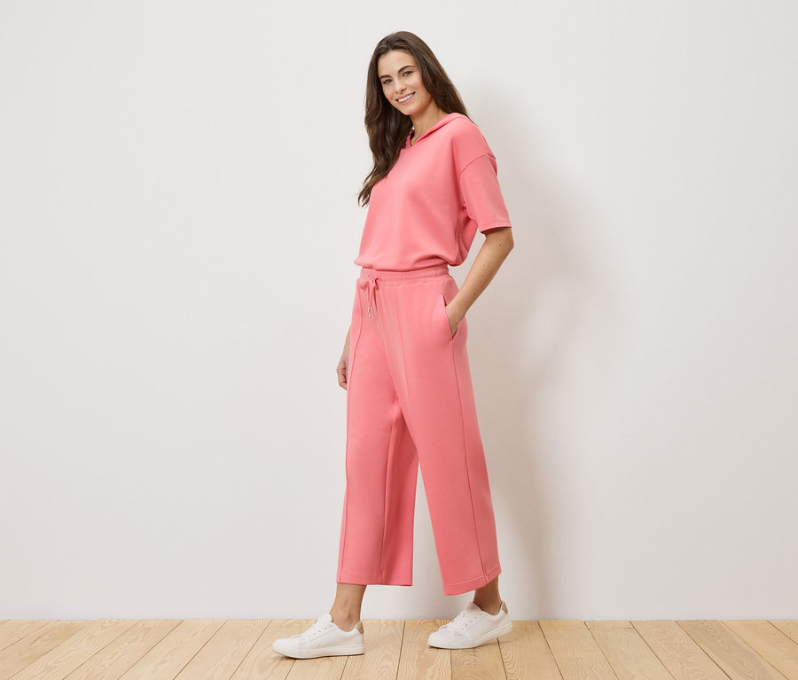 Une femme porte un survêtement corail avec un haut à capuche et une culotte de jupe, ainsi que des baskets blanches.