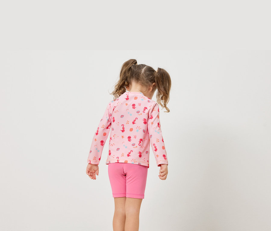 Une petite fille avec des tresses porte un t-shirt rose à manches longues avec des motifs marins et un short rose.