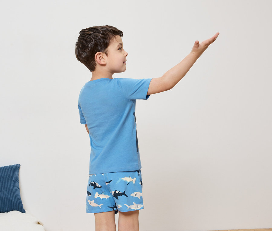 Un garçon aux cheveux bruns, vêtu d'un t-shirt bleu clair et d'un short à motifs de requins, se tient de côté, le bras levé.