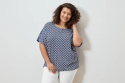Femme souriante aux cheveux bouclés portant un chemisier bleu et blanc et un pantalon blanc.