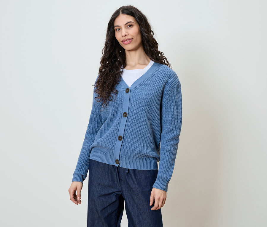 Femme aux longs cheveux bouclés et foncés portant un cardigan bleu et un jean foncé.