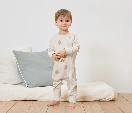 Un jeune enfant se tient pieds nus devant un lit avec des coussins et tient une chaîne en bois. Il porte une combinaison claire à motif oursons avec fermeture éclair.