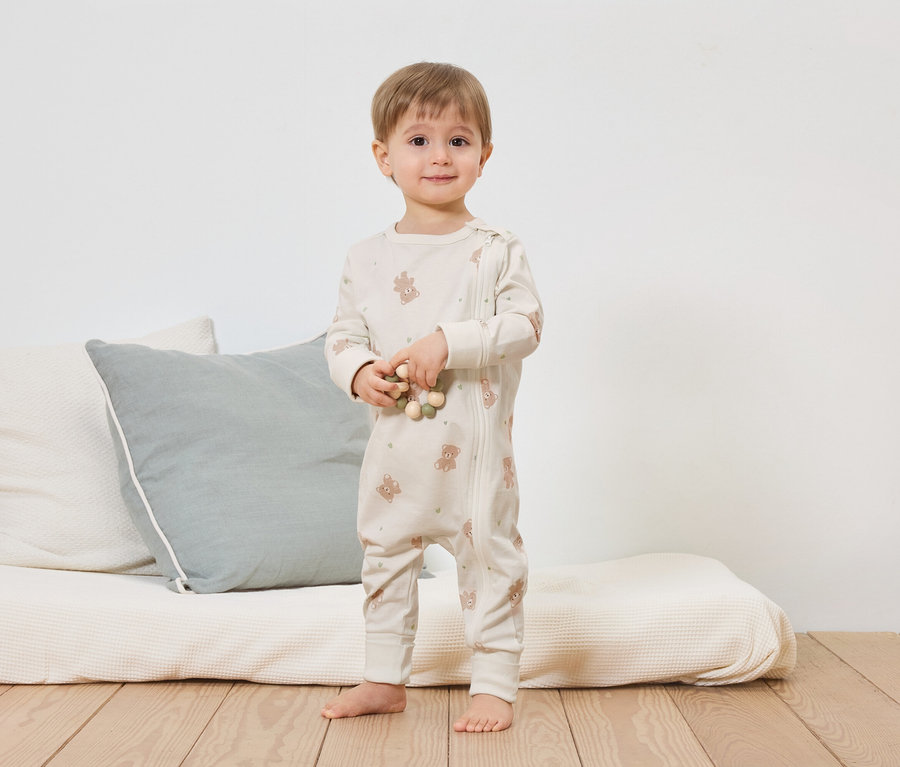 Un jeune enfant se tient pieds nus devant un lit avec des coussins et tient une chaîne en bois. Il porte une combinaison claire à motif oursons avec fermeture éclair.
