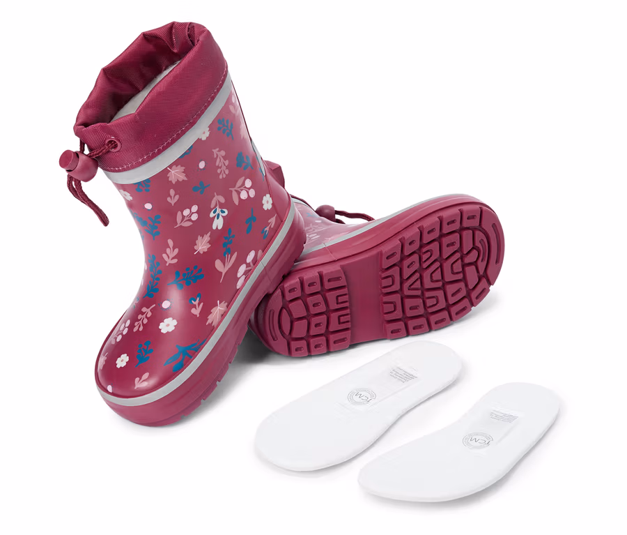 Une paire de bottes en caoutchouc bordeaux à motifs floraux et deux semelles intérieures blanches sont posées sur un fond blanc.