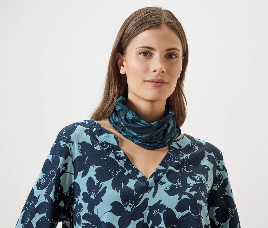 Une femme porte un haut bleu à motifs floraux et un cache-cou assorti sur un fond blanc.