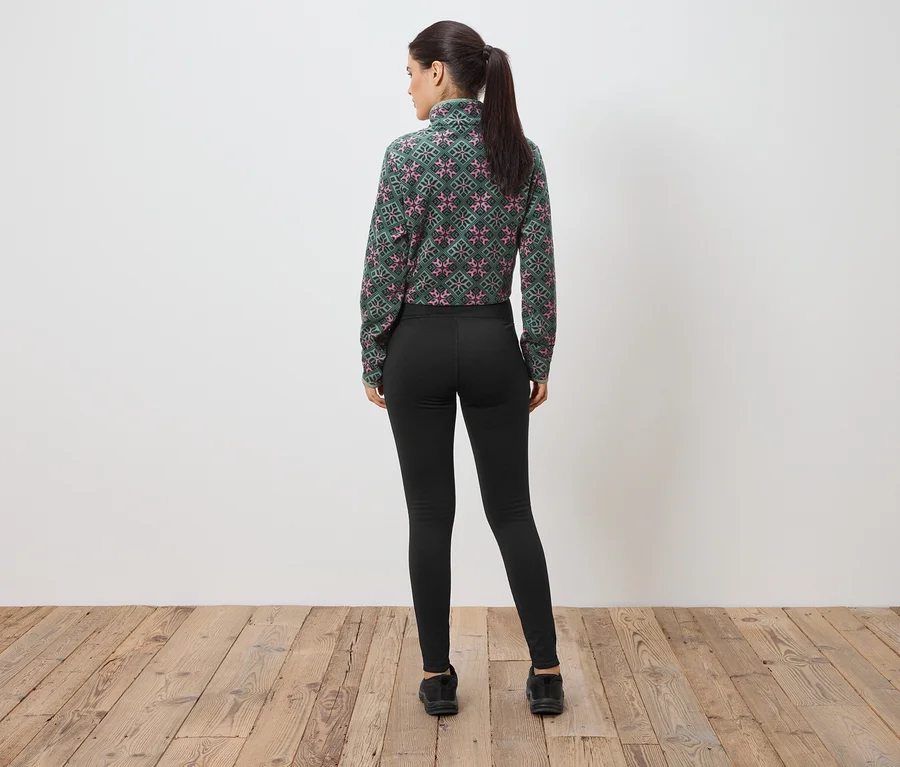 Femme vue de dos portant un chemisier à motifs vert et rose, un pantalon noir et des chaussures noires devant un mur.