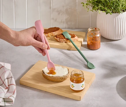 Une main étale de la confiture sur un pain avec une spatule rose. À côté se trouvent une spatule verte et deux pots de confiture.