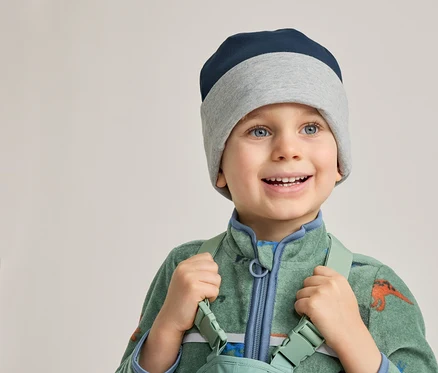 Un garçon souriant portant un bonnet bleu et gris et une combinaison verte à motifs de dinosaures tient des bretelles.