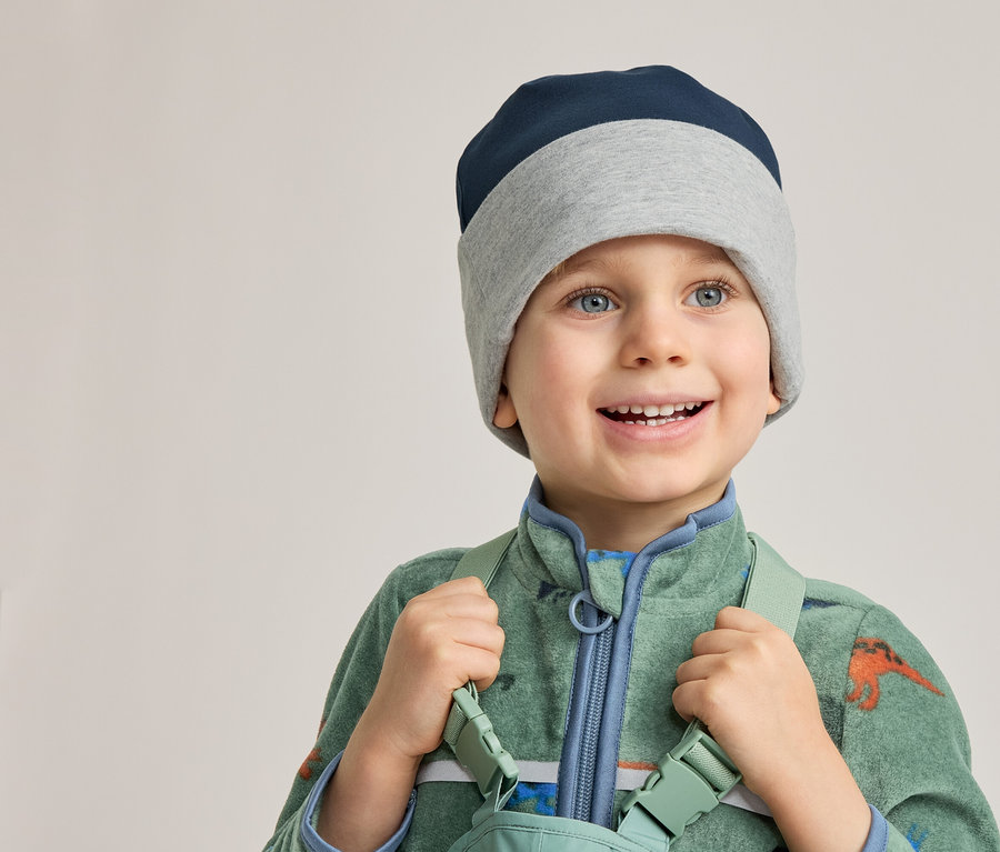 Un garçon souriant portant un bonnet bleu et gris et une combinaison verte à motifs de dinosaures tient des bretelles.