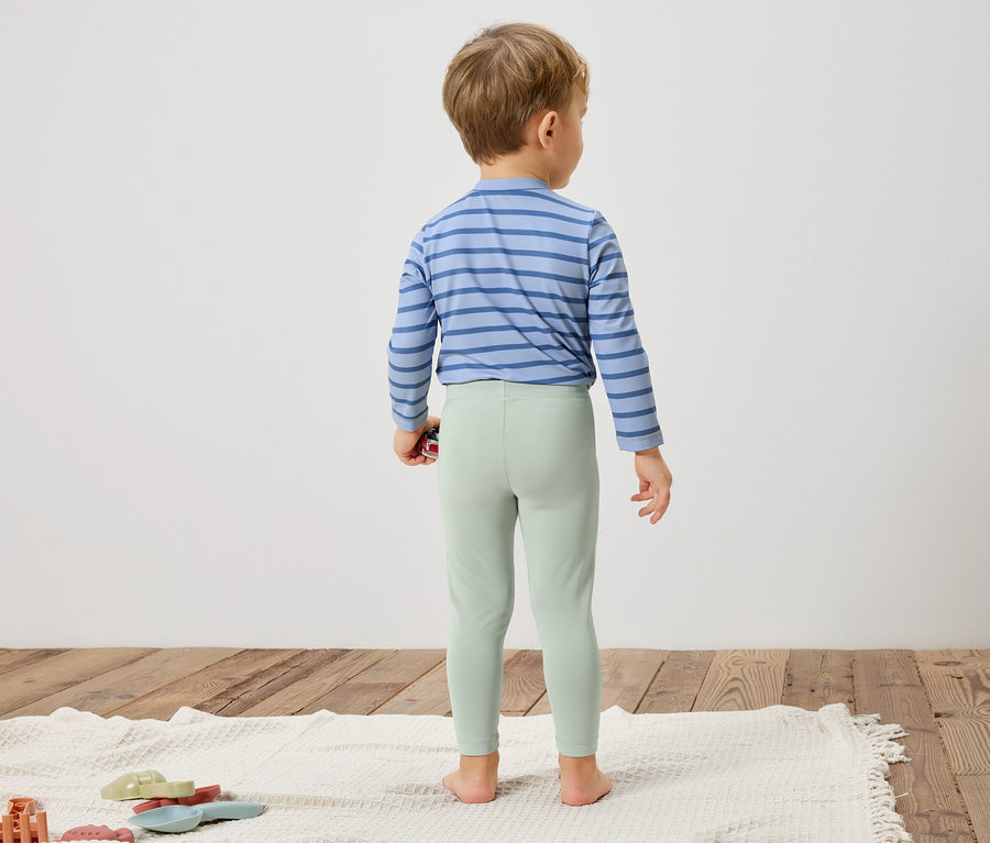 Un enfant se tient dos à la caméra, portant une chemise à rayures bleues et des leggings vert clair.
