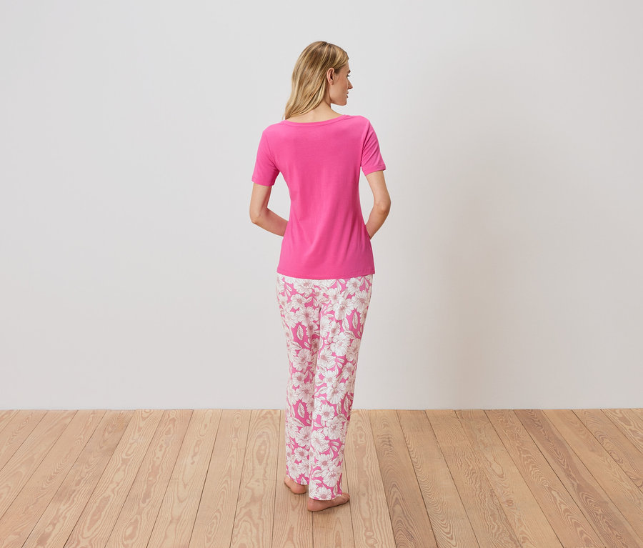 Vue arrière d'une femme blonde portant un t-shirt rose et un pantalon de pyjama rose à motifs floraux, debout sur un plancher en bois.