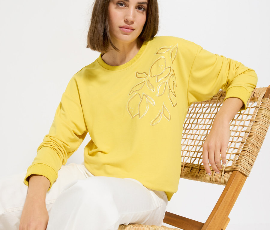 Femme assise sur une chaise portant un pull jaune avec motif de citron et un pantalon blanc.
