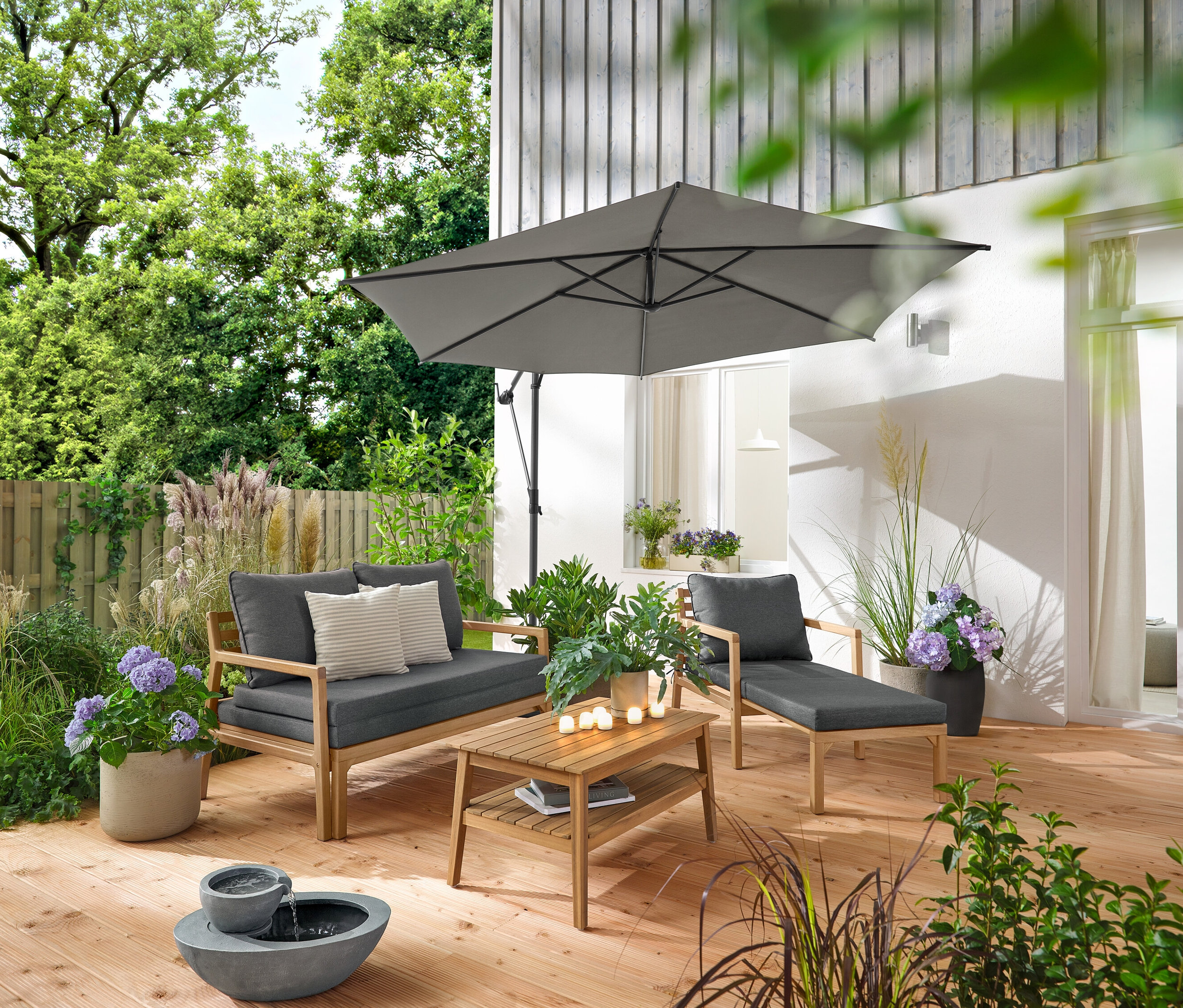 Sur une terrasse en bois se trouvent un fauteuil de jardin extensible, un canapé de jardin extensible, une table basse de jardin d'environ 80 x 50 cm et une fontaine de jardin solaire. Un parasol Leco anthracite crée de l'ombre. 2 coussins d’extérieur avec texture lin et 6 bougies LED rechargeables d’extérieur décorent l'espace.
