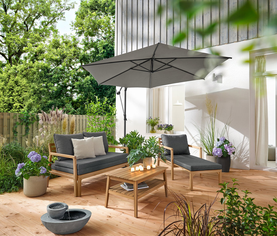 Sur une terrasse en bois se trouvent un fauteuil de jardin extensible, un canapé de jardin extensible, une table basse de jardin d'environ 80 x 50 cm et une fontaine de jardin solaire. Un parasol Leco anthracite crée de l'ombre. 2 coussins d’extérieur avec texture lin et 6 bougies LED rechargeables d’extérieur décorent l'espace.