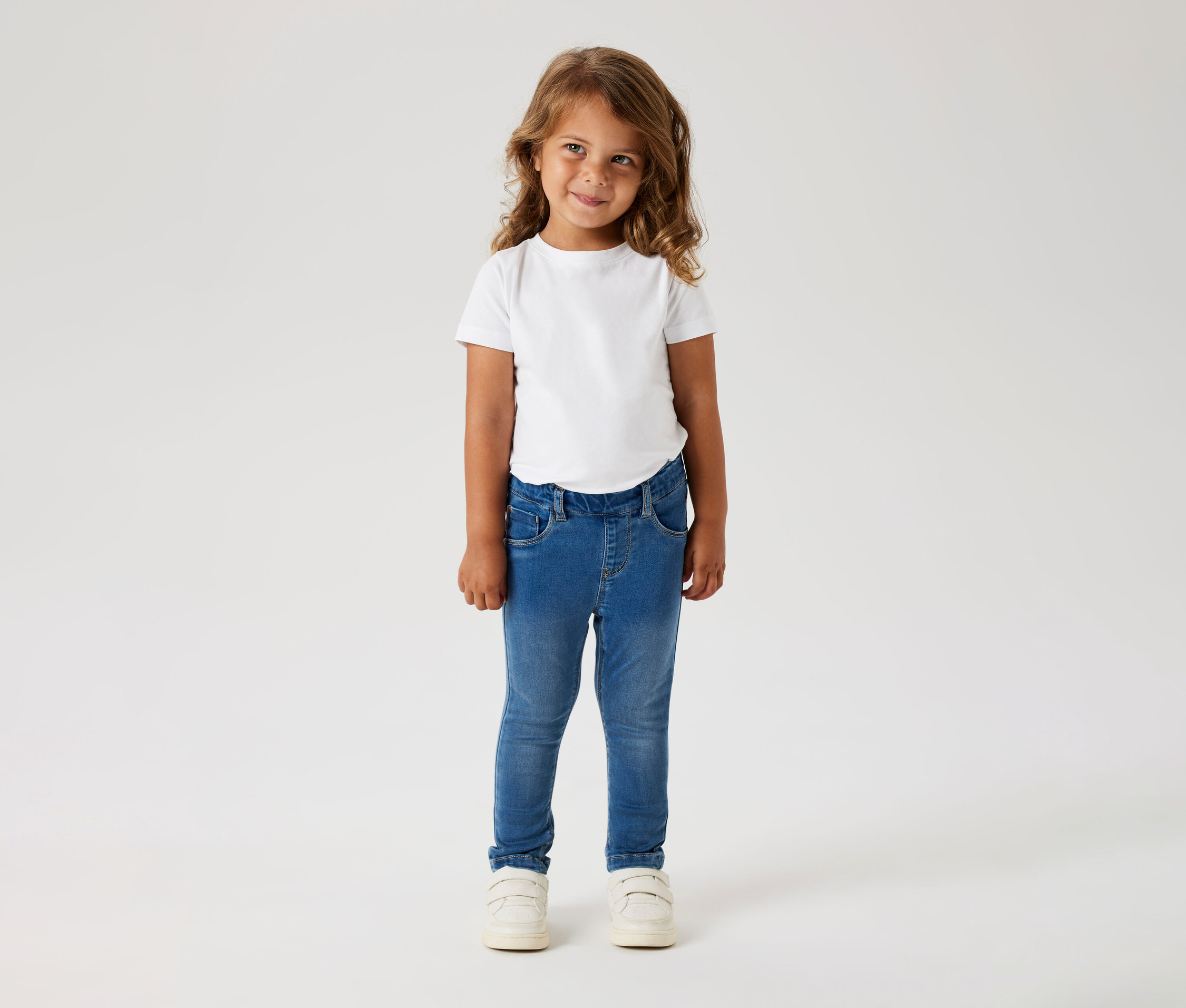 Fillette aux cheveux bouclés portant un t-shirt blanc, un jean bleu et des chaussures blanches.