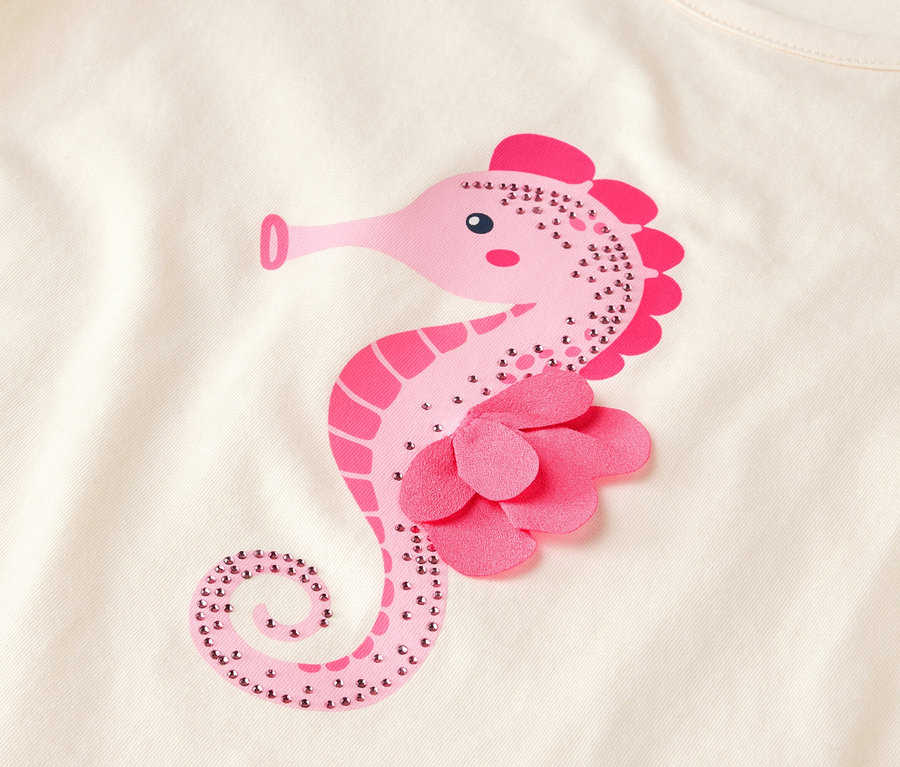 Gros plan d'un tissu de couleur claire avec un motif d'hippocampe rose orné de strass et d'une fleur en tissu.