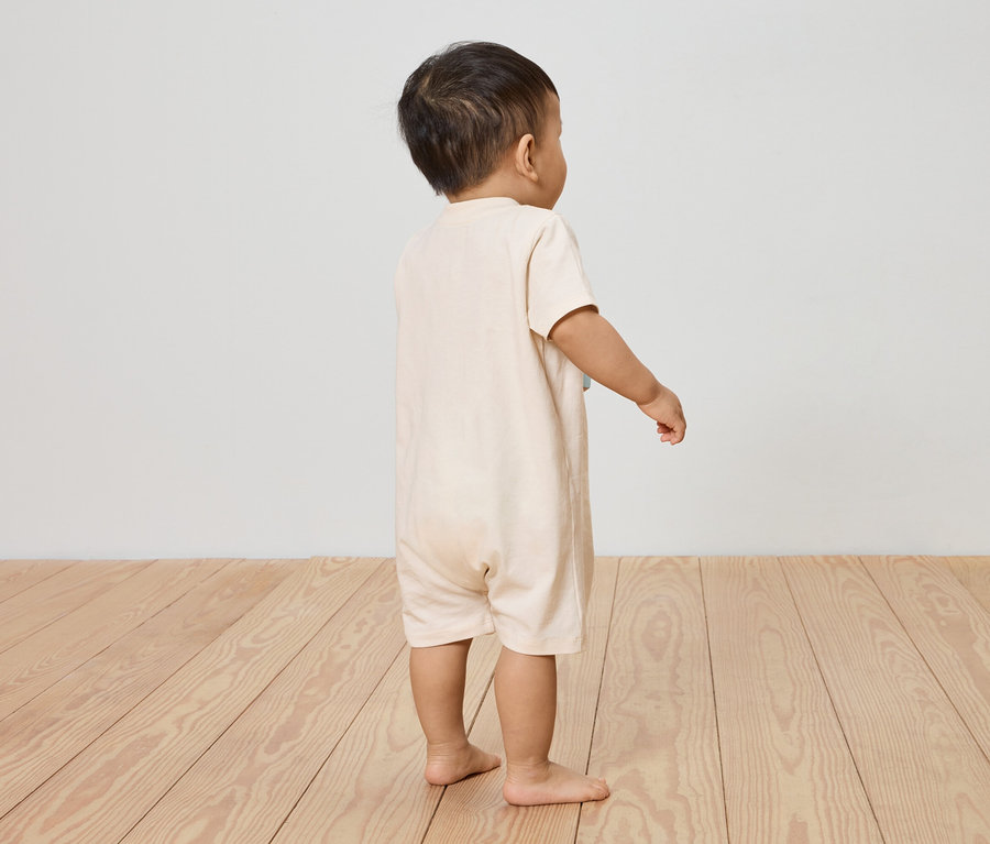 Un enfant se tient dos à la caméra sur un plancher en bois, portant une combinaison à manches courtes beige clair.