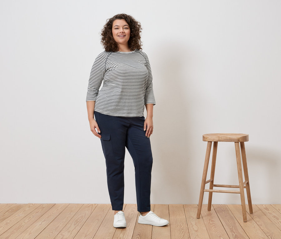 Une femme aux cheveux bouclés se tient debout en portant un haut rayé, un pantalon bleu et des baskets blanches à côté d'un tabouret.