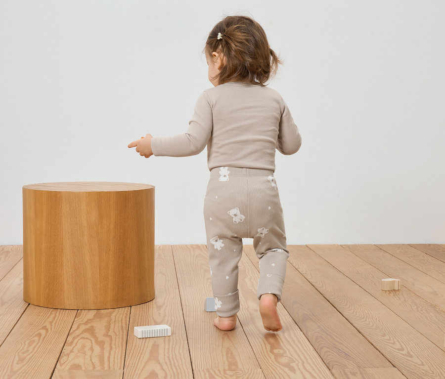 Un enfant en bas âge, vêtu de vêtements beiges à motifs d'ours, pointe du doigt un tabouret en bois et des jouets sur un plancher en bois.