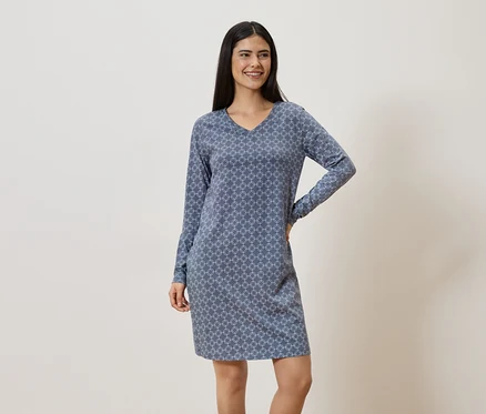 Une femme souriante aux cheveux foncés porte une robe bleue à motifs à manches longues.