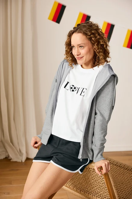 Femme portant un short en molleton, un t-shirt de sport sur le thème du football et une veste molletonnée grise assise devant une guirlande allemande.