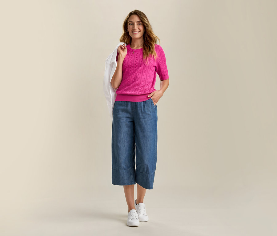 Une femme souriante porte une veste en jean et un pull en maille fine rose avec des torsades. Elle porte une culotte bleue et des baskets blanches.