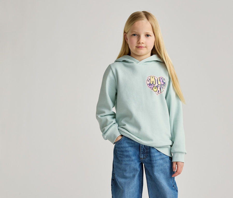 Une fille blonde porte un sweat à capuche bleu clair avec un imprimé cœur et un jean.