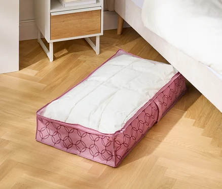 Sur un plancher en bois, un sac de rangement rose est placé sous un lit à côté d'une table de chevet.